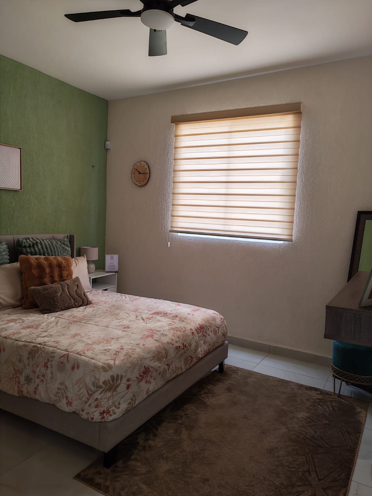 Recámara con pared de acento en verde, cama con edredón floral, buró con lámpara y cómoda con espejo